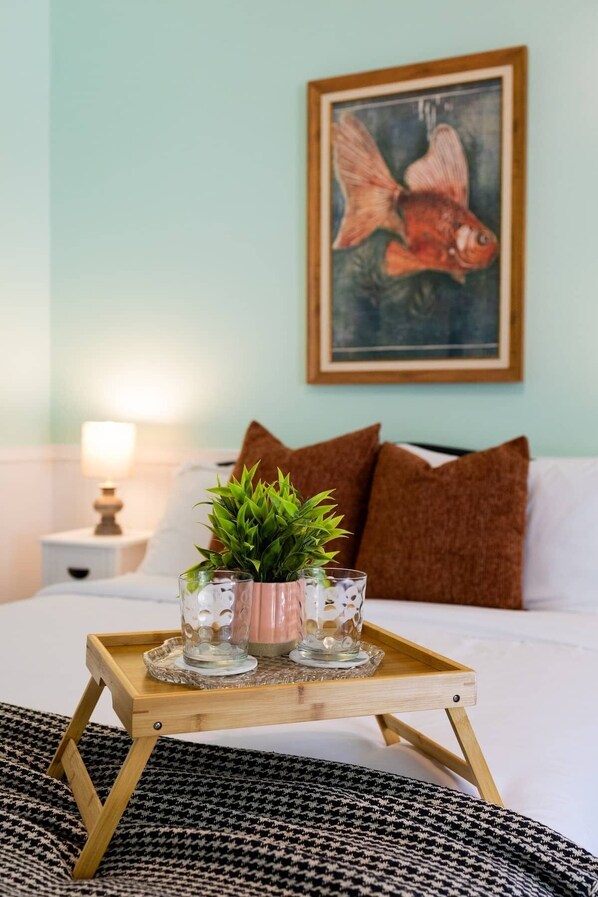 Ein Bett mit einem hölzernen Tablett, das Glasvasen und eine Pflanze trägt, ein Fischgemälde an der Wand und ein Nachttisch mit einer Lampe.