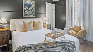 A bedroom with a bed, bedside tables, a nightstand, a chair, a window with curtains, and a painting on the wall. - Flower Room | Team Joseph Ellen (St. Augustine)