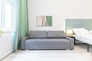 A modern bedroom with a grey sofa, a bed with white linens, a bedside table with a lamp, and a green curtain.