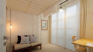 A cozy room with a wooden ceiling, a beige sofa with patterned cushions, a wooden dining table with a chair, and a large window with sheer curtains. - TWH Hostel Bangkok (Bangkok)