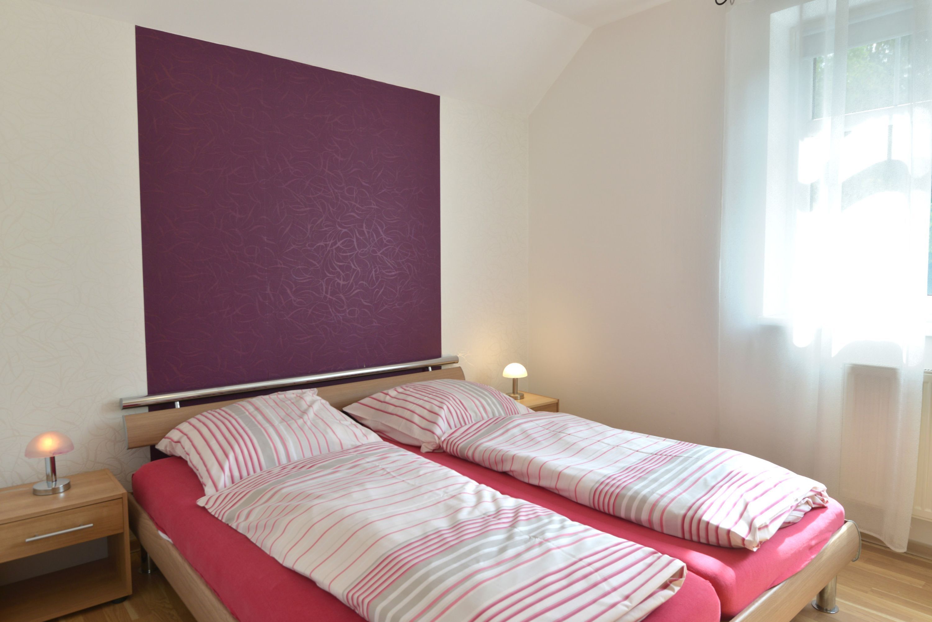 Un lit double avec une literie à rayures dans une chambre dont un grand mur est peint dans une nuance de violet, une table de chevet avec une lampe et une fenêtre ornée de rideaux légers.