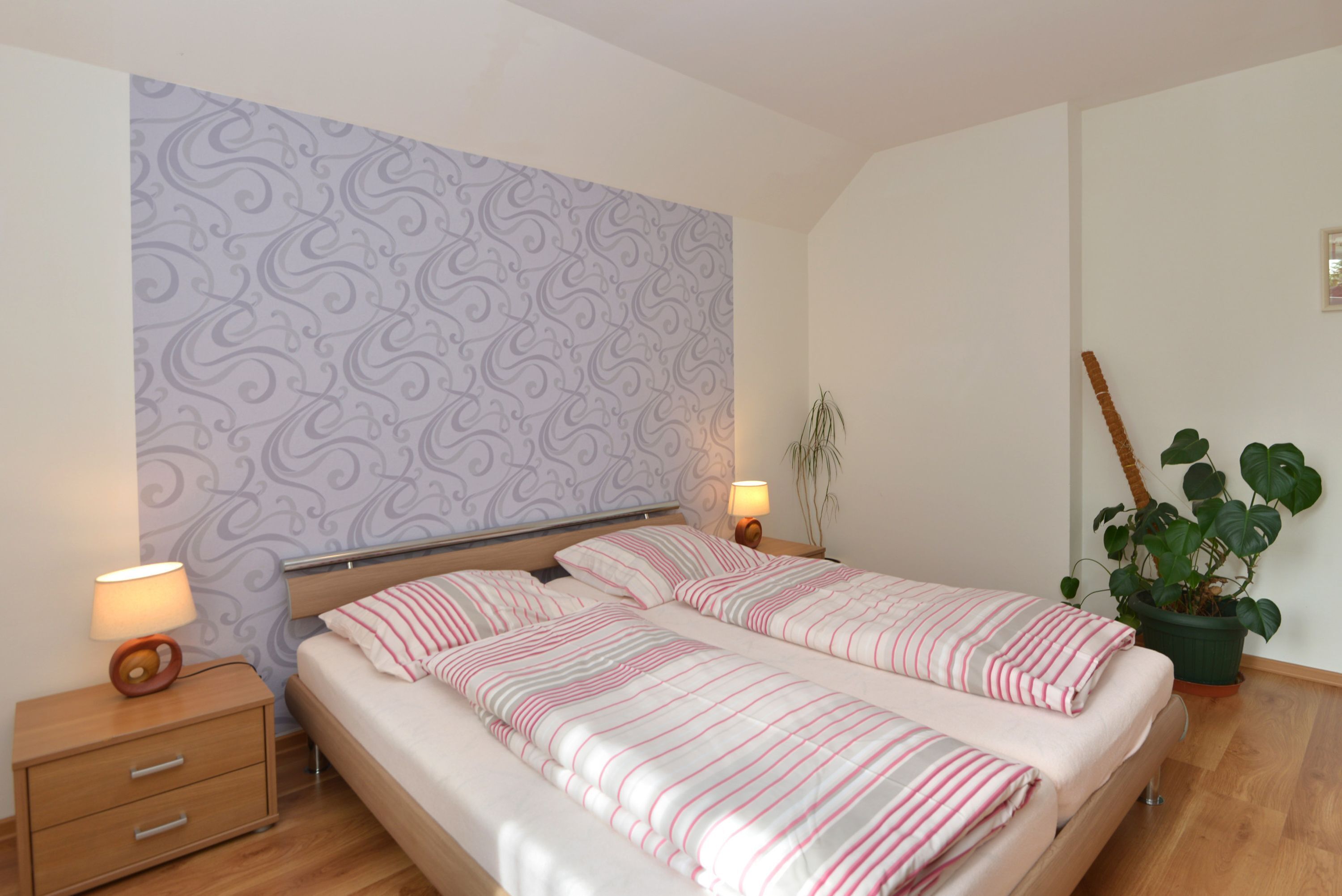A double bed with striped bedding, a nightstand with a lamp, a potted plant, and a patterned wallpaper on the wall.