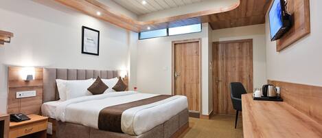 A modern hotel room with a large bed, wooden headboard, bedside table with a phone, a desk with a kettle, and a TV mounted on the wall.