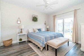 Quarto com cama, mesas de cabeceira, um banquinho, ventoinha no teto e vista para o exterior.