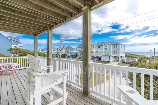 A wooden deck with white railings, a bench, and a chair. In the background, there are houses with a view of the ocean.