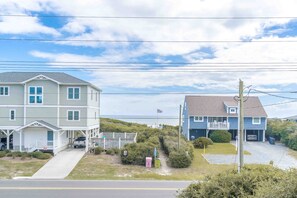 A coastal residential area with two houses, one with a porch and the other with a balcony, and a clear view of the ocean. - Seas The Moment - 2nd Row, 4 BR Home with Covered Porches (Surf City)