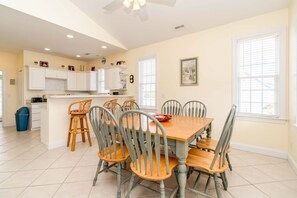 A dining area with a wooden table and six chairs, a kitchen with white cabinets, and a trash bin. - Seas The Moment - 2nd Row, 4 BR Home with Covered Porches (Surf City)