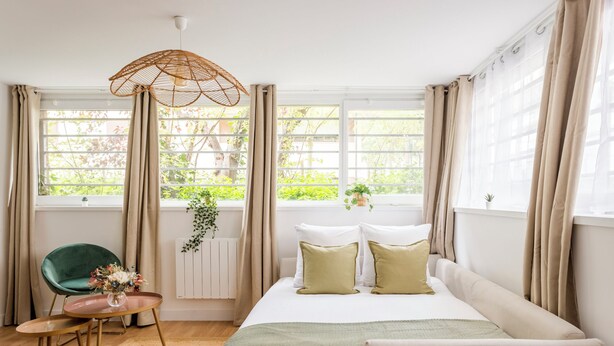 A bedroom with a bed, a green chair, a small table, and a hanging plant. - Family Suite - Marais - Pelee (Paris)