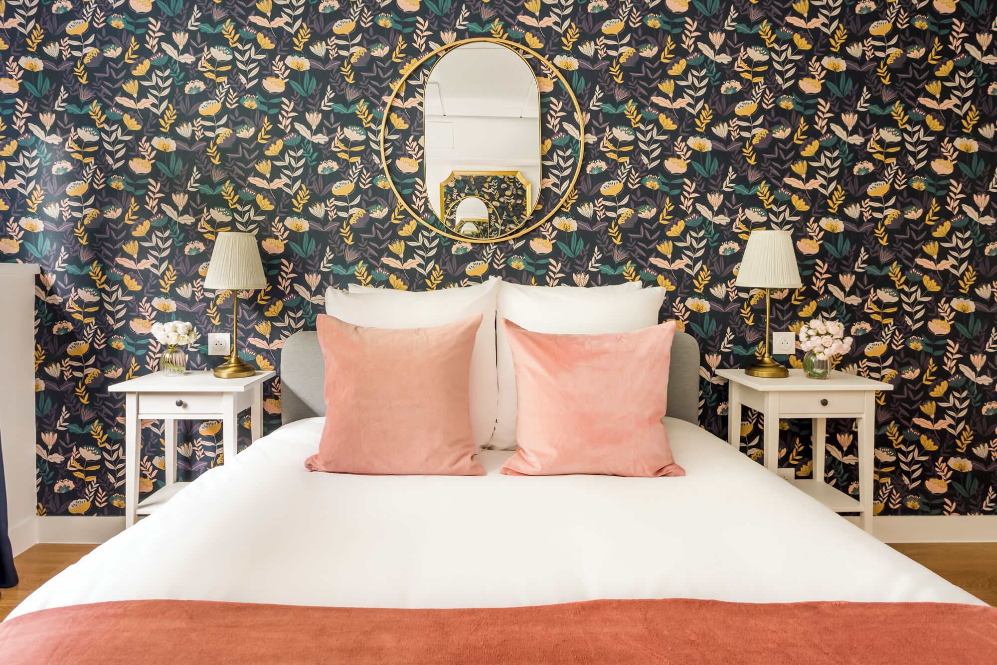 Une chambre avec un papier peint à motifs floraux, un grand lit avec des coussins roses, deux tables de chevet avec des lampes et un miroir au mur.
