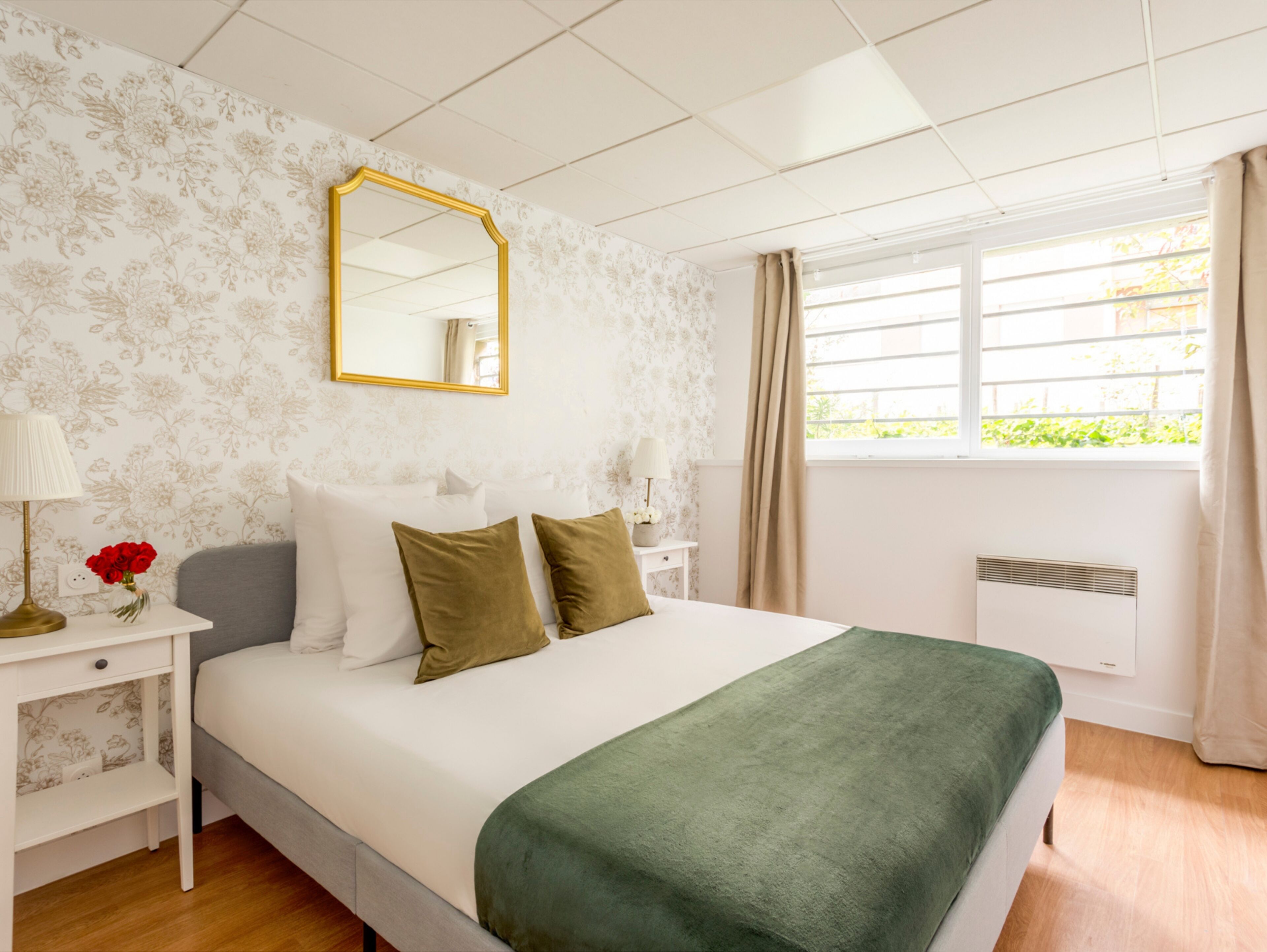 Un dormitorio ordenado con cama, mesita de noche, espejo y ventana con cortinas.