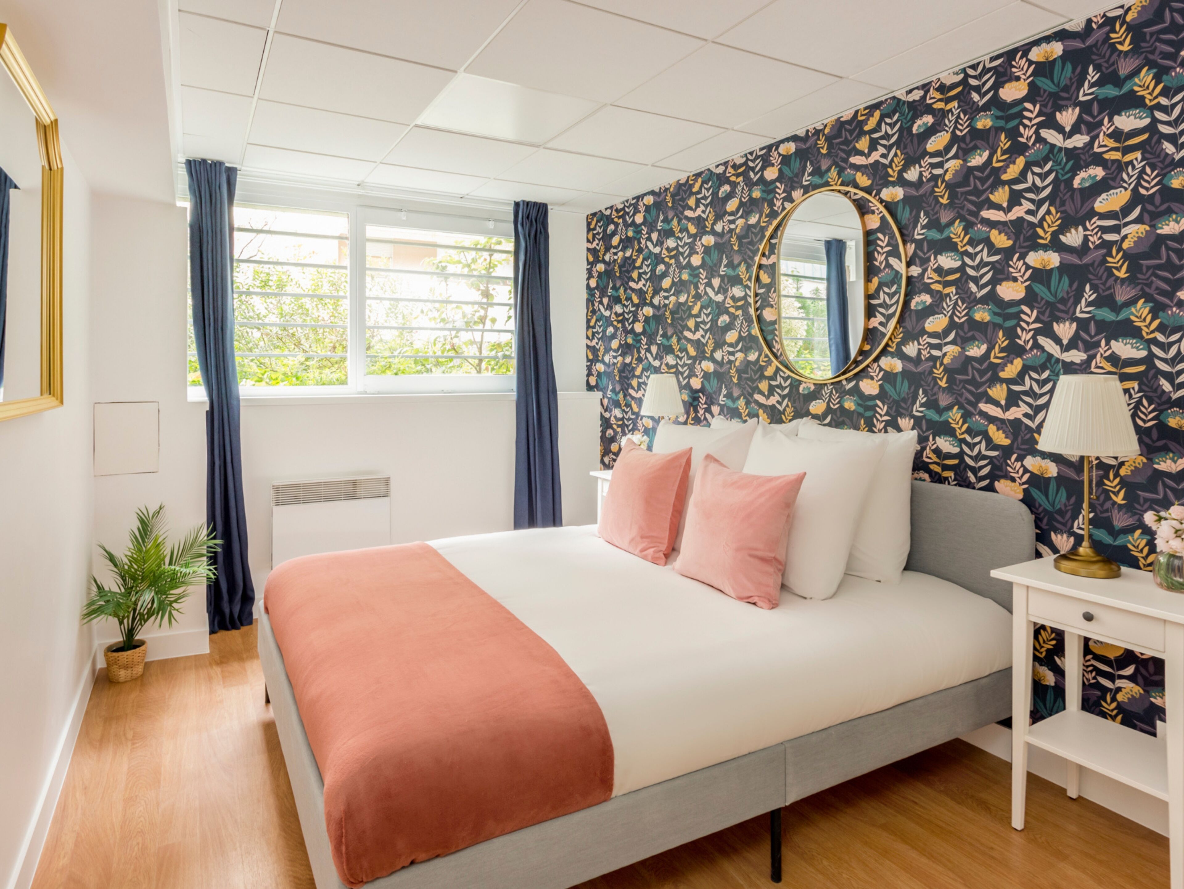 Un dormitorio moderno con una cama grande, papel tapiz floral, un espejo redondo y una mesita de noche con una lámpara.