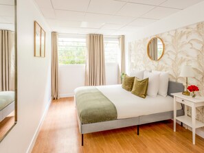 Moderni hotellihuone, jossa on suuri sänky, yöpöytä lampun kanssa ja näkymä vehreään maisemaan ikkunasta.