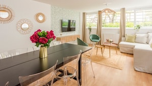 A modern living room with a dining area, a TV mounted on the wall, and a sofa with a view of greenery outside. - Family Suite - Marais - Pelee (Paris)