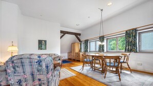 Una sala de estar con sofá, una mesa de comedor con sillas, vigas de madera en el techo y una ventana con cortinas.