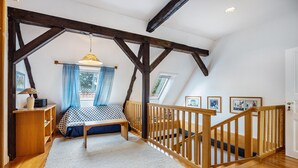 Un dormitorio con techo inclinado, vigas de madera, una cama con colcha de estrellas azules y blancas, un escritorio de madera con lámpara y una escalera con barandilla de madera.