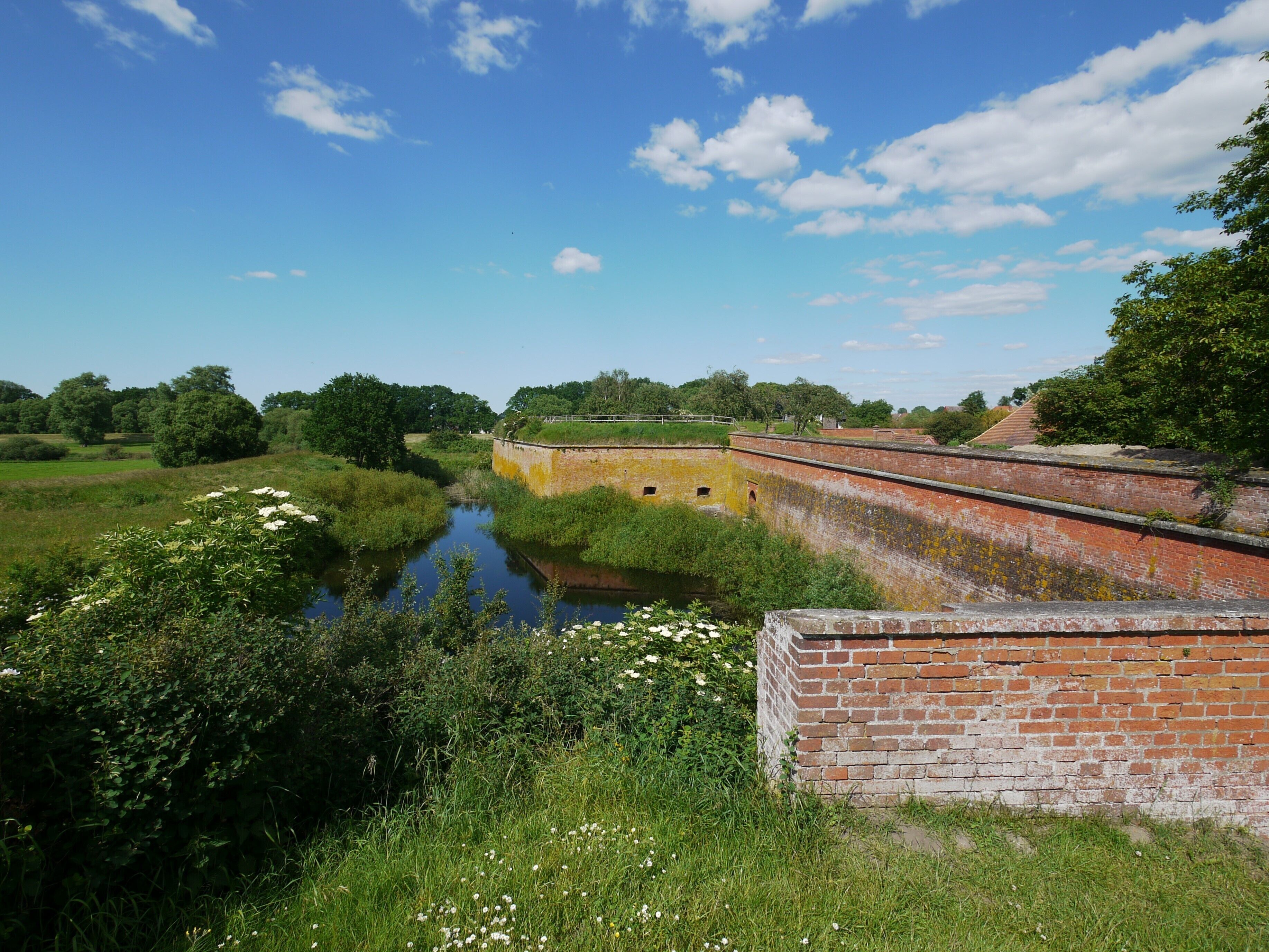 A historic brick structure with a river in the foreground, surrounded by greenery and trees.