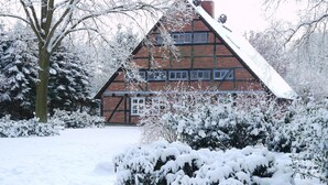 Una casa de ladrillo tradicional con un techo empinado, rodeada de árboles y arbustos cubiertos de nieve.