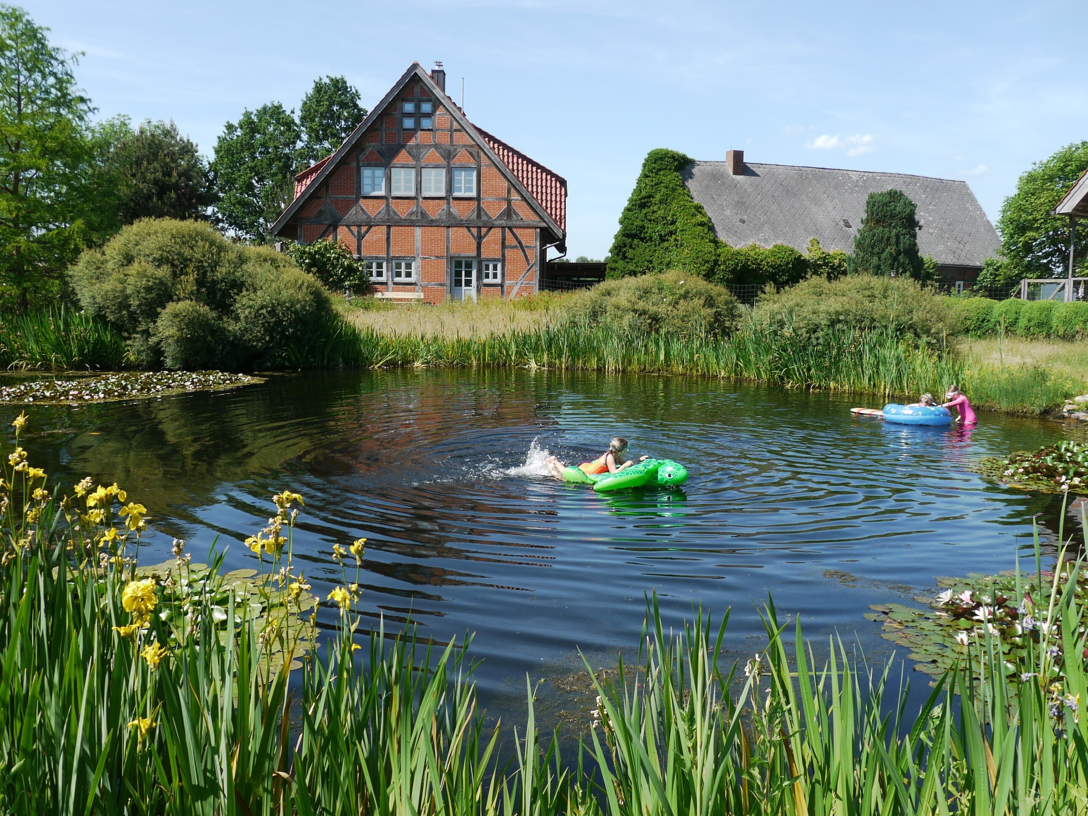 A calm river with two people on inflatable rafts, surrounded by lush greenery and a traditional house.