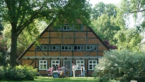 En traditionel bindingsværksbygning med en græsplæne foran, et spiseområde med borde og stole, og et stort træ der giver skygge.