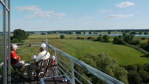 En terrasse med udsigt over sø og marker, med gelændinger i metal og et bord med stole.