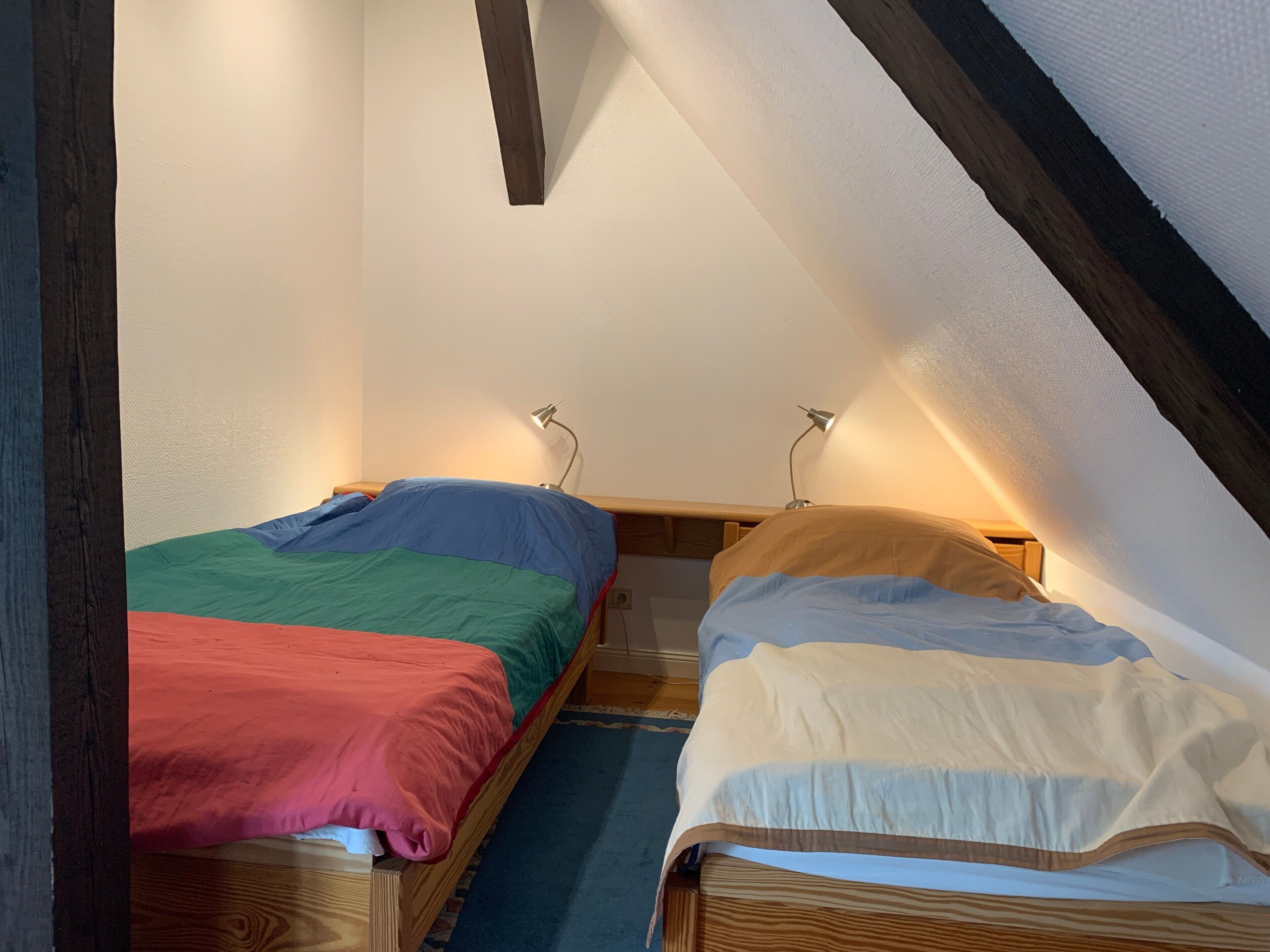 Une petite chambre mansardée, chaleureuse et accueillante, avec deux lits en bois, un plafond en pente et une seule lampe fixée au mur.