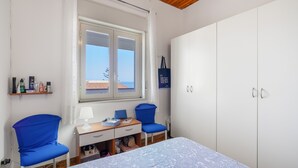 A bedroom with a bed, two blue chairs, a desk with a lamp, a wardrobe, and a window with a view of the sea.
