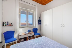 A bedroom with a bed, two blue chairs, a desk with a lamp, a wardrobe, and a window with a view of the sea.