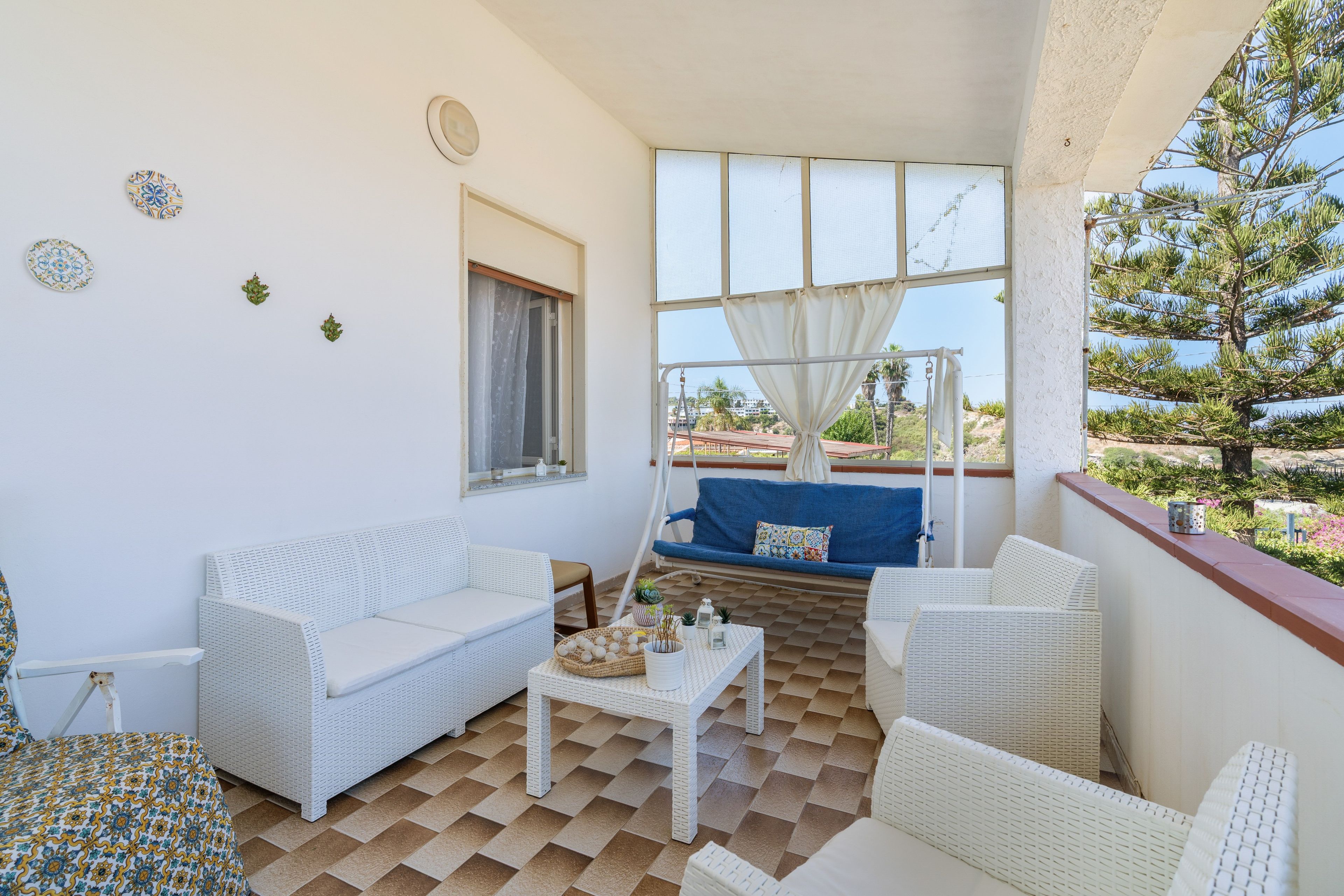 Ein Balkon mit weißen Korbmöbeln, einem kleinen Tisch mit Dekoration und Blick auf einen Baum sowie entfernte Gebäude.