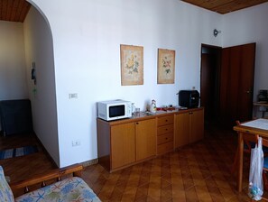 Cocina compacta con microondas, cafetera y un refrigerador pequeño. Hay armarios de madera y piso de baldosas.