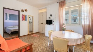 A compact living space with a bed, a dining table with chairs, a TV, and a window with curtains. - Casa Orchidea with Veranda – Private Terrace, Wi-Fi & Air Conditioning (Costa Saracena - Castelluccio)