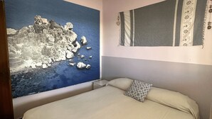A bedroom with a bed, a nightstand, a wall-mounted lamp, and a large painting of a coastal landscape. - Casa Orchidea with Veranda – Private Terrace, Wi-Fi & Air Conditioning (Costa Saracena - Castelluccio)