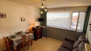 Una sala de estar acogedora con comedor, un sofá, una mesa para dos personas, una lámpara, un radiador y una ventana con cortinas.