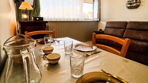 Una mesa de comedor dispuesta con platos, vasos y una jarrón, con un sofá marrón y una ventana al fondo.