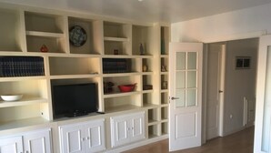 Una sala de estar con televisor, estantes con diversos objetos y una puerta que da a otra habitación.