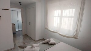 Una habitación de hotel con cama, ventana con cortinas traslúcidas y un pasillo que conduce a otra habitación.