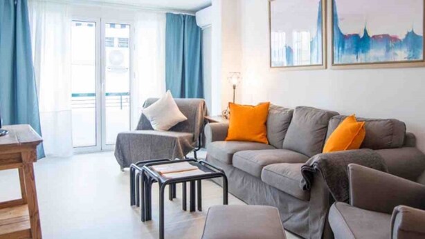 A modern living room with a grey sofa, orange cushions, and a wooden coffee table. There is a chair with a white cushion and a lamp on a side table. - Comfortable apartment near Seville city center (Sevilla)