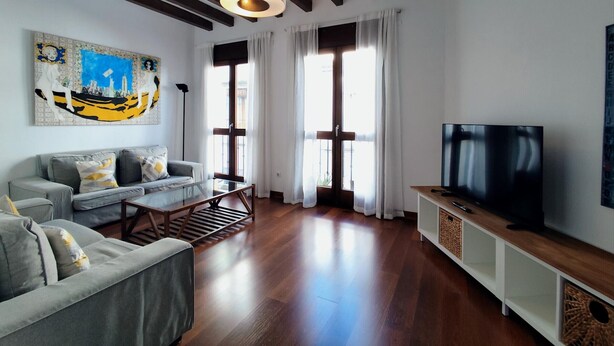 A modern living room with a sofa, a coffee table, a TV on a wooden stand, and a large window with curtains.