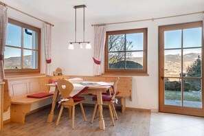 Un comedor con un banco, mesa y sillas de madera, ventanas amplias con cortinas y vistas al exterior.