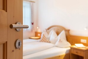 A wooden door with a keyhole and a handle, leading to a bed with white bedding and pillows, a bedside table, and a nightstand with a lamp.