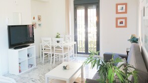 Ein Wohnzimmer mit Fernseher, weißen Möbeln, einer Couch, einem Tisch und einer Topfpflanze.