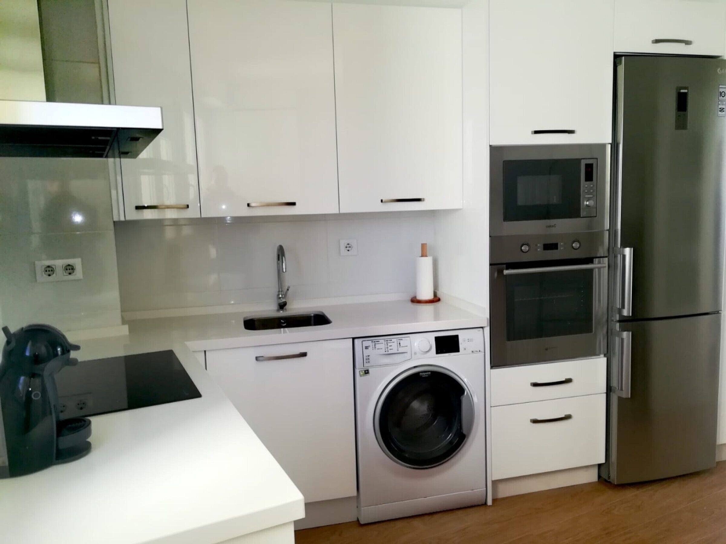 Een moderne keuken met witte kasten, een roestvrijstalen koelkast, een inbouwoven, een magnetron, een wasmachine en een koffiezetapparaat.