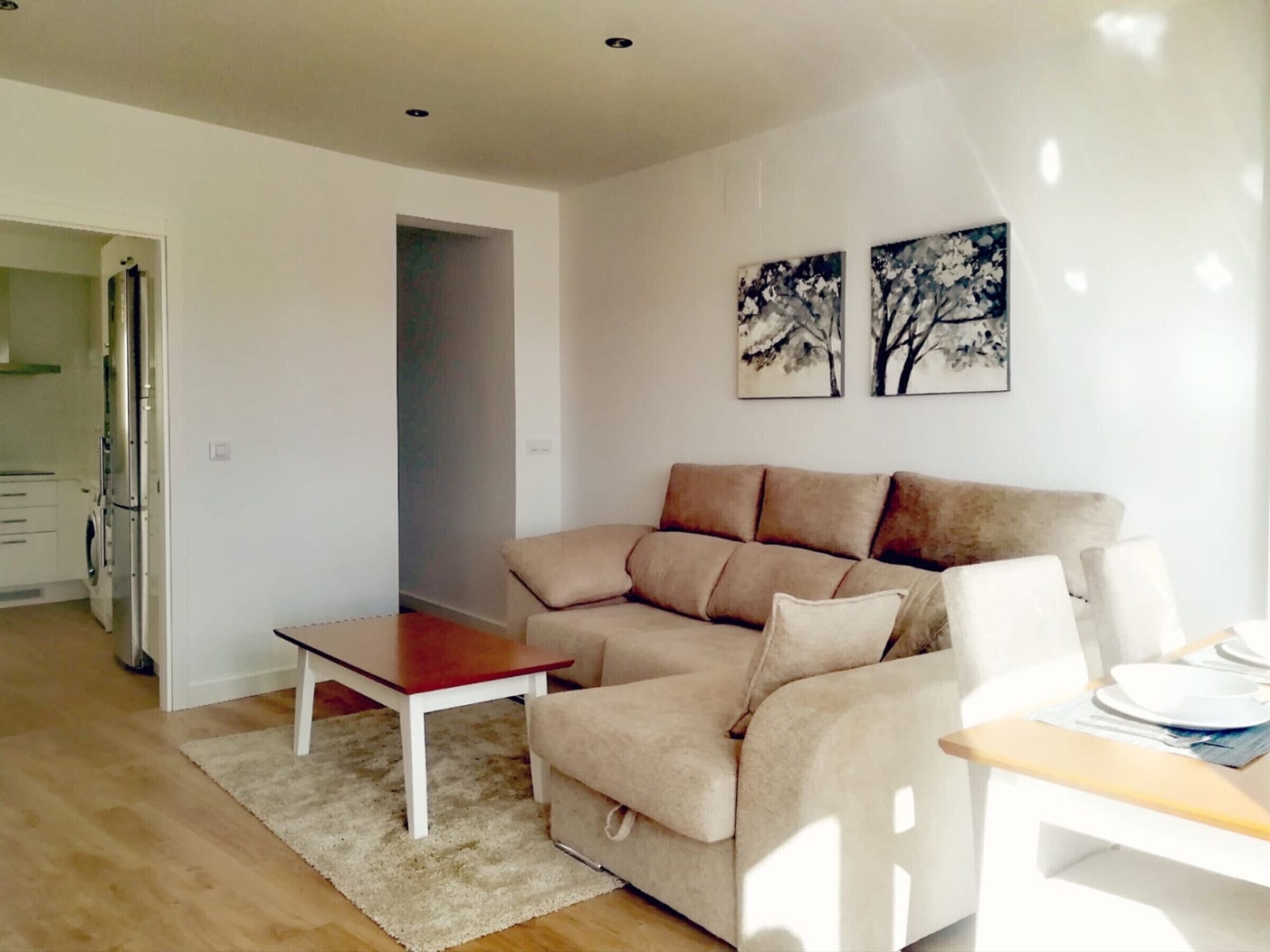 Een moderne woonkamer met een beige bank, een houten salontafel, en een eethoek met een witte tafel en stoelen. Er hangen twee schilderijen van bomen aan de muur.