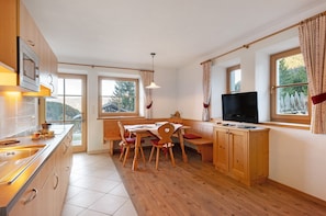 A kitchen with wooden cabinets, a built-in microwave, a dining table with chairs, a TV, and large windows with curtains.