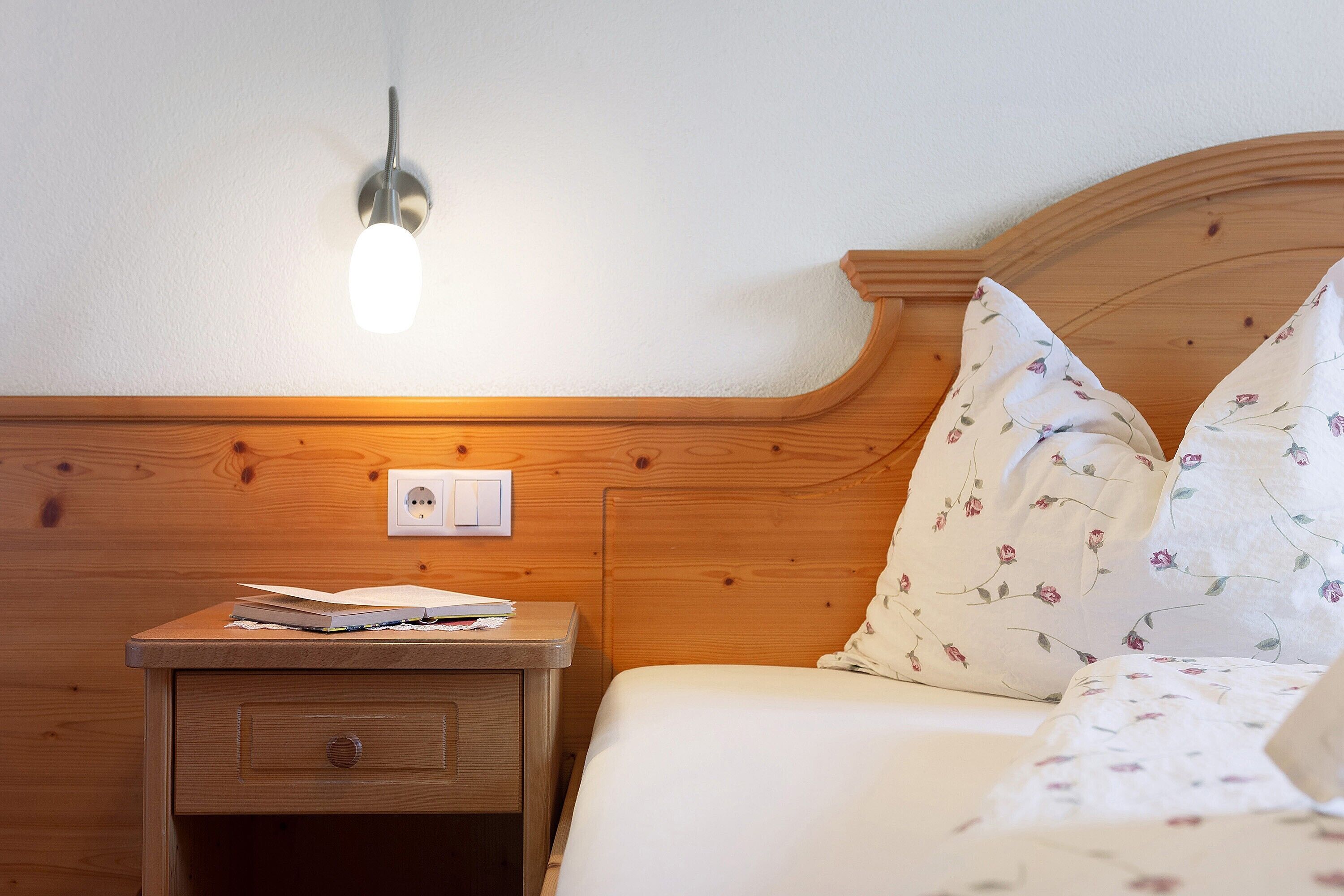 Una mesita de noche de madera con una lámpara, una cama con ropa de cama estampada con motivos florales y un enchufe de pared.
