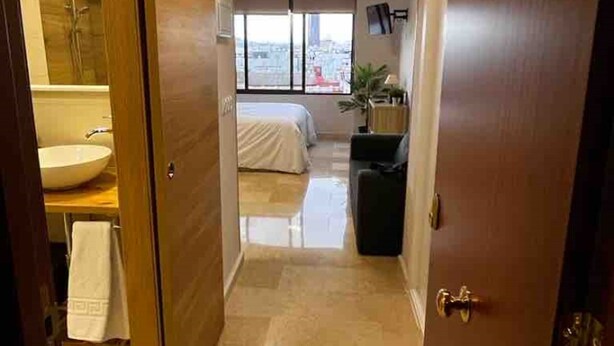 Habitación de hotel con ventana grande, cama, mesita de noche y baño con lavamanos y espejo.