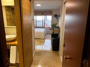 A hotel room with a large window, a bed, a bedside table, and a bathroom with a sink and mirror. - Elegant Triana apartment with views, near Seville center (Sevilla)