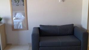 Una habitación con un sofá de color gris oscuro, un mueble de madera, un espejo que refleja una cama con sábanas blancas y un termostato fijado a la pared.
