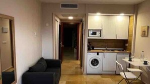 Una habitación compacta con cama, un pequeño sofá, una kitchenette con microondas y lavadora, y un comedor con mesa y sillas.