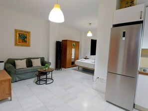 A modern living room with a sofa, a small round table with plants, a wooden cabinet, and a kitchen area with a stainless steel refrigerator. - Peaceful studio for couples in Seville center (Sevilla)
