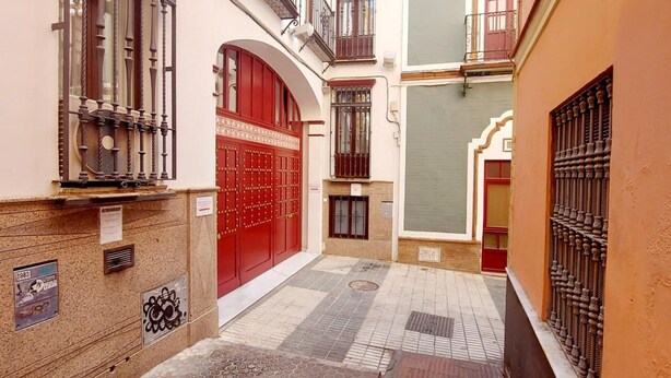 A narrow alleyway with a red gate, a tiled pavement, and a building with a balcony.
